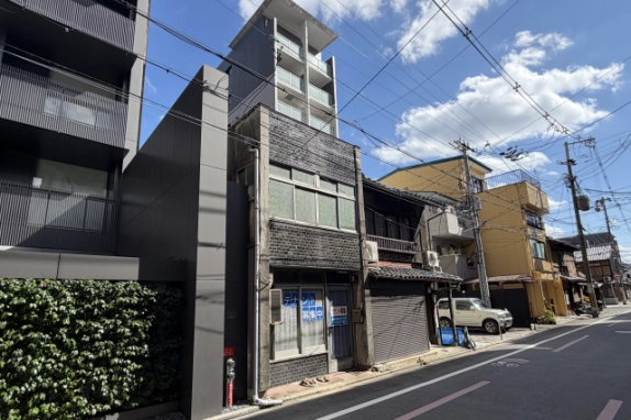 小川通沿いに建つ店舗を左斜め前から見た外観。左に植栽のある灰色の建物、右に瓦の庇とシャッターの町家が隣り合う