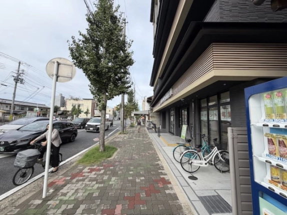 建物を右手に見た歩道と車道。街路樹の奥へ続く道と店舗前の自転車