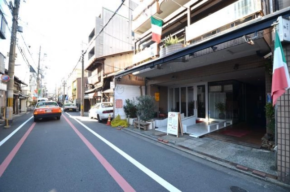 通りの奥へ延びる道路と、右側に建つビルの1階。ガラス張りの店舗前に白いウッドデッキと植栽が並ぶ