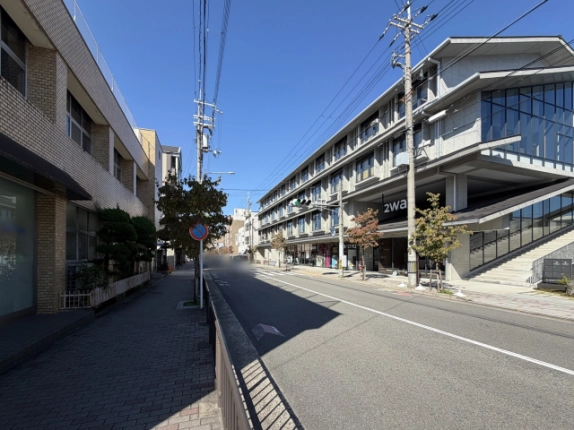 向かいの歩道側から見た前面道路。右手に2waの建物と屋外階段、道路沿いに街路樹と電柱が並ぶ
