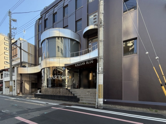 右斜め前から見た外観。ダークブラウンの外壁に銀色の曲線状の庇と丸いガラス窓が並び、右手前に電柱が立つ