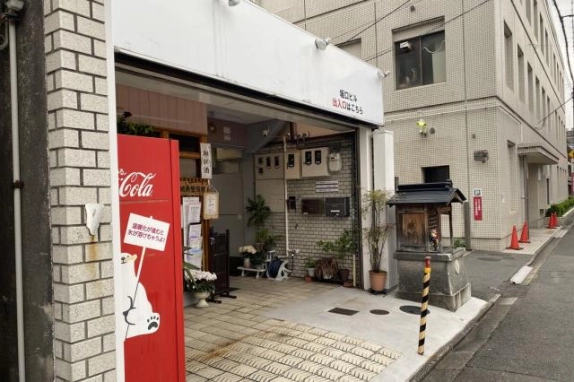 左斜めから見た坂口ビルの共用出入口。タイル敷きの通路の奥に白いレンガ調の壁とメーターボックス、鉢植えが並び、右手に小さな祠がある