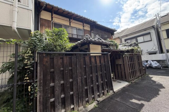 路地から斜めに見た外観。濃い色の板塀と左側の植栽越しに、2階の窓と瓦屋根、玄関の小屋根が見える