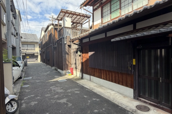 町家の玄関前から延びる西陣の路地。右手に格子窓と玄関が続き、左手には車やバイクが停まっている