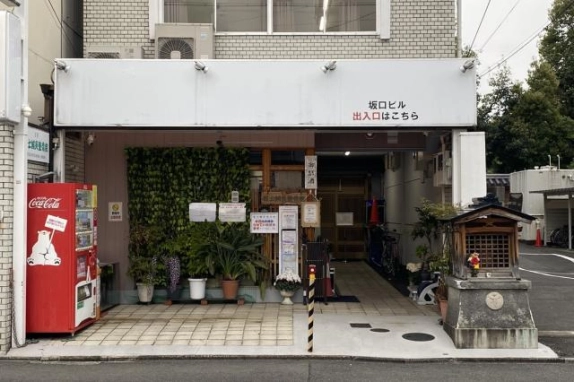 1階エントランスの正面。白い看板の下、左に赤い自動販売機と緑の壁面や鉢植え、中央に奥へ続く通路、右に小さな祠が並ぶ