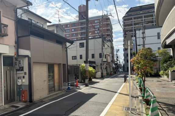 上京区の通り沿いで、2階が貸事務所・店舗区画となる建物を左手に見た眺め。右側に黄色い舗装の歩道と街路樹、奥に中高層マンションが並ぶ