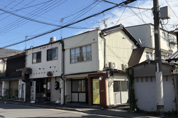 通り沿いに右斜め前から見た建物。側面の室外機と1階の黄色いガラス戸、右手に電柱とシャッター付きの小屋が見える