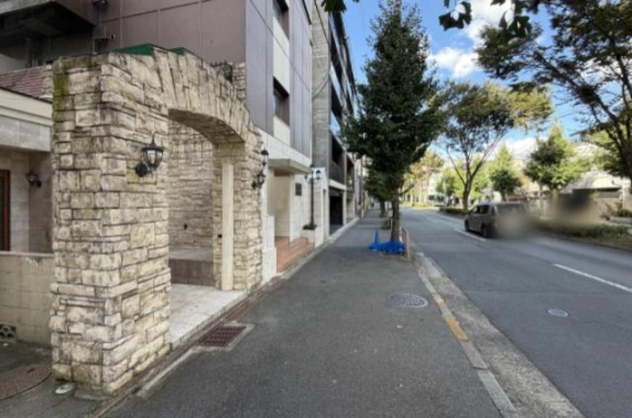 石積みのアーチと壁付けランタンがある建物前の歩道。右側には街路樹と車道が奥まで続く
