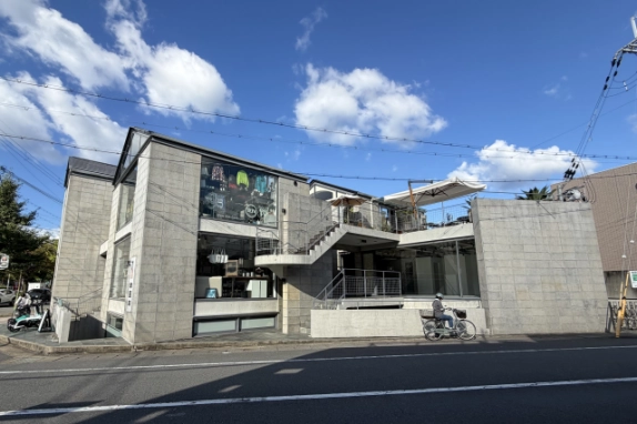 道路の向かいから見たデザイナーズビルの正面。上下2段のガラス開口と、屋上テラスへ上がるコンクリートの外階段、白いパラソル