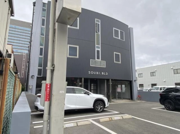 左斜め前から電柱越しに見たビル外観。建物前の駐車場に白い車が停まり、左側に細長い窓が縦に並ぶ