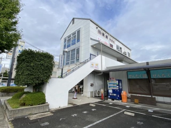建物の側面に沿って2階へ延びる白い外階段。階段下に自動販売機、右隣にせんたく館、手前に駐車区画