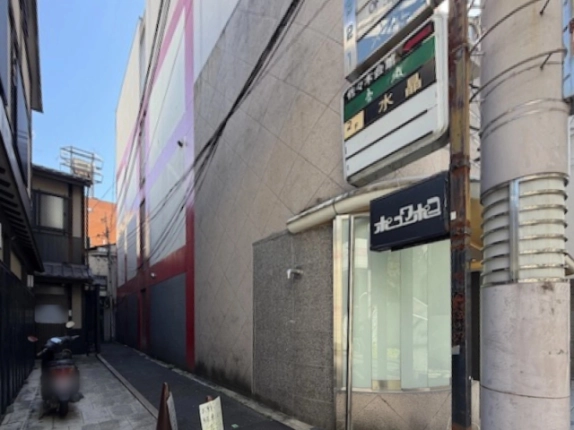 東山区祇園の通りから見た路地の入口。左に町家と停めたバイク、正面に赤と紫のラインが入る建物、右手前に看板と電柱