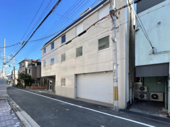 ビルを右斜め前から見た外観。1階に大きな白いシャッター、左奥にガラス扉の入口、手前に電柱、右隣の建物の前に室外機が並ぶ