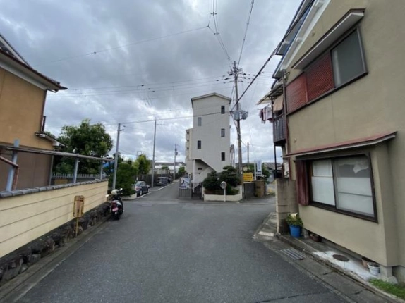 住宅に挟まれた道路の突き当たりに見える白い建物。左手に塀とバイク、右手に2階建て住宅がある