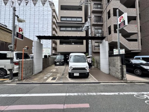 道路の向かいから見た正面の門構え。白い軽自動車の右のタイル通路を傘を差した人が歩き、左右にコインパーキングの看板が立つ