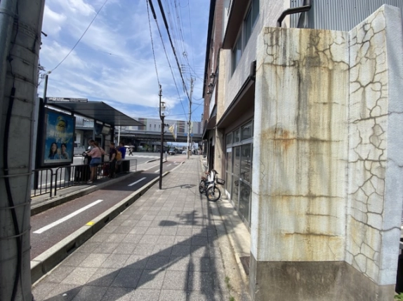 店舗を右手に見た前面歩道。左手に屋根付きのバス停、奥に高架が見える