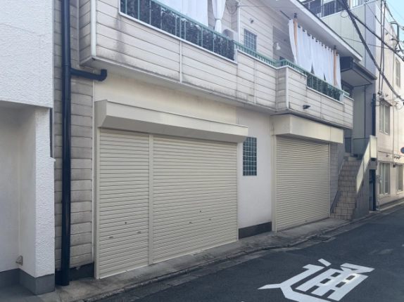 左斜め前から見た外観。黒い縦樋の右に庇付きのシャッターが2か所続き、建物の右端に外階段が見える