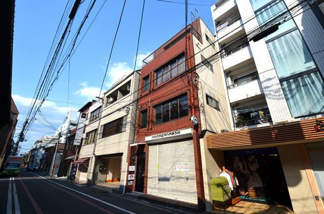 FORCE TOMINOKOJIを右斜め前から見上げた外観。赤レンガ調タイルの建物で、1階にシャッターと黒い入口ドアがあり、右隣に白いビルが並ぶ