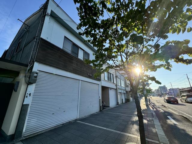 七条通大宮西入の木造2階建一棟貸店舗事務所を斜め下から見上げた外観。白い外壁に木目調の帯、1階に2連の白いシャッター、右手に街路樹越しの日差しと車道