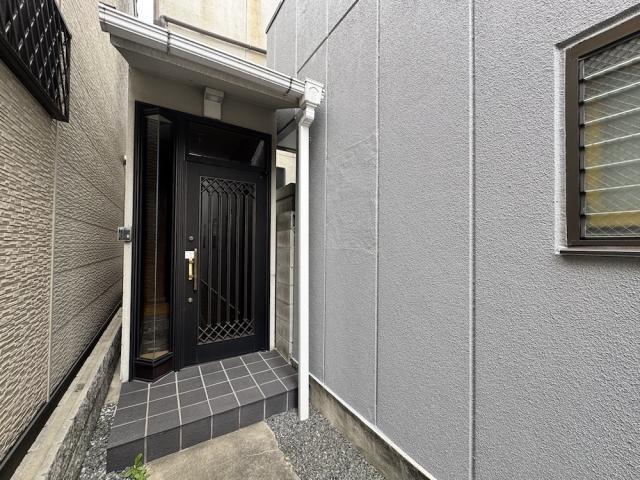 戸建の玄関まわり。格子模様の黒い玄関ドアとタイル張りの段差、白い柱の小さな庇があり、右側にルーバー窓のあるグレーの壁