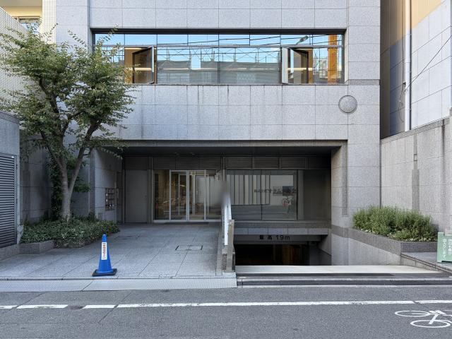 新町アイエスビルの正面外観。グレーの石張り外壁、植栽の奥にガラスの自動ドアのエントランス、右手に高さ制限1.9mの地下への車路