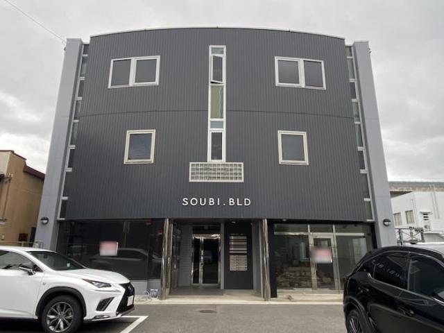 伏見区竹田藁屋町のSOUBI BLDを正面から見た外観。濃いグレーの外壁に「SOUBI.BLD」の文字があり、1階は中央の入口を挟んで左右がガラス張り