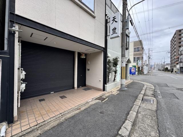 右京区西京極南庄境町の1階テナントを右斜め前から見た外観。黒いシャッターとタイル張りの入口前があり、右手に歩道と道路が続く