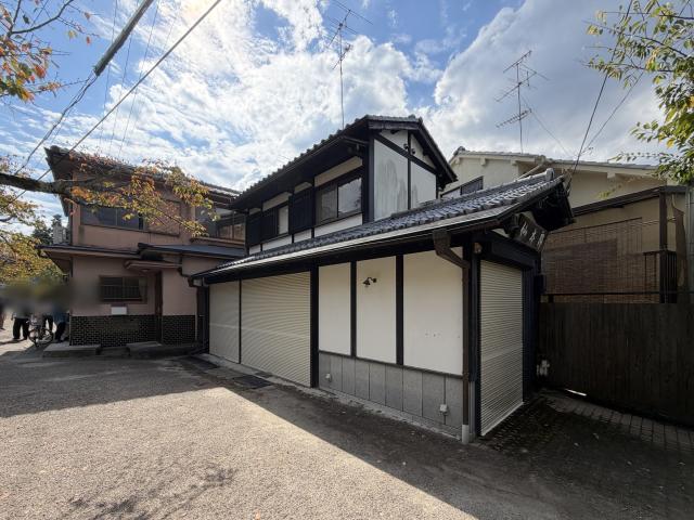 左京区の下南田町店舗の外観を斜め前から見た様子。瓦屋根に白壁と黒い木部の2階建てで、1階にベージュのシャッターが並び、手前に砂利敷きの地面が広がる